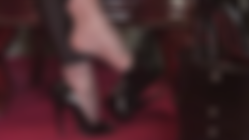 Nahaufnahme der füße der herrin, an denen glänzende lackpumps mit offener spitze in superglänzenden grauen strumpfhosen baumeln.