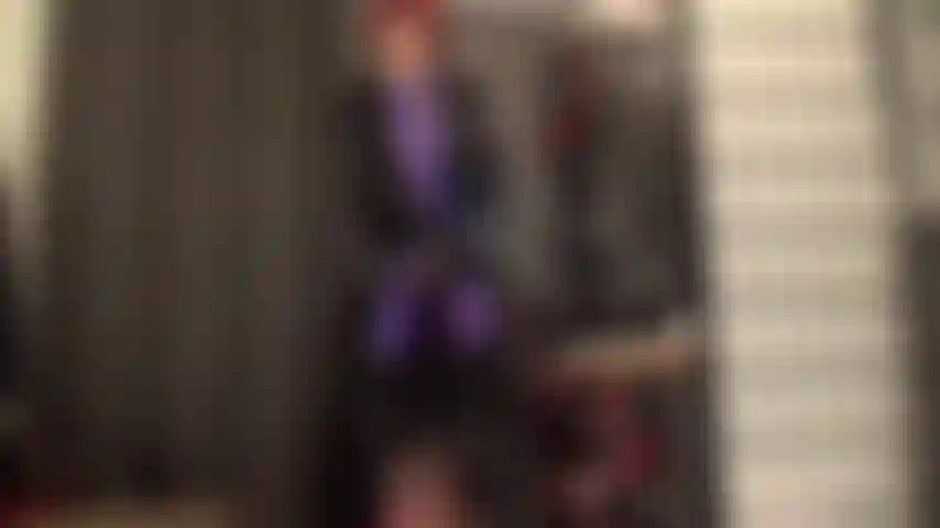 Ein glänzender, schwerer pvc-mantel über einem lila leohex-anzug mit stryker-strapon und stiefeln.